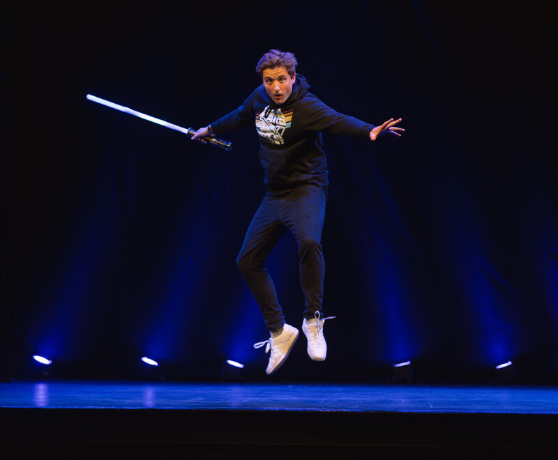 Comedian Jon Hipkiss leaping in the air wearing a Star Wars t-shirt and holding a light sabre.