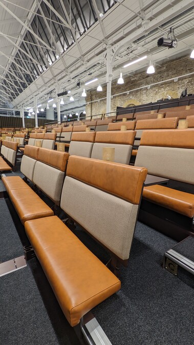A photo of one of our seating banks, taken from part way up the steps looking diagonally across several rows. The seats are padded, leather bench seats, with tables and lamps, and are a burnt orange colour with a light beige seat back. The high glass ceiling, metal framework and hanging lights of the Fireworks Factory is visible in the background.