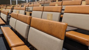 A photo of one of our seating banks, taken from part way up the steps looking diagonally across several rows. The seats are padded, leather bench seats, with tables and lamps, and are a burnt orange colour with a light beige seat back. The high glass ceiling, metal framework and hanging lights  of the Fireworks Factory is visible in the background.