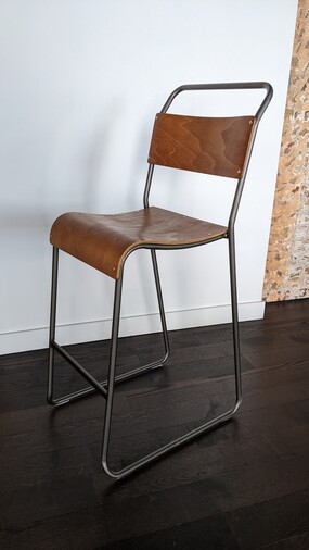 A photo of a tall stool, with a wooden seat and back rest, and a metal frame, with a foot rest.