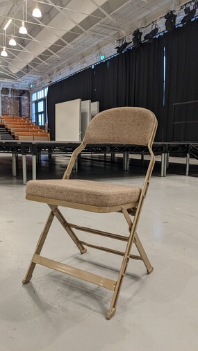 A photo of a loose folding chair that is comfortably cushioned with a padded seat and back. The chair is light beige in colour and has a metal frame and legs.
