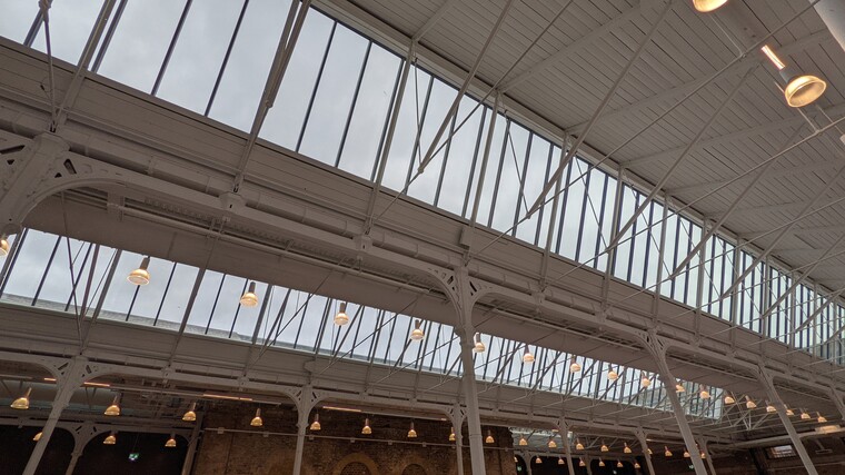 Exposed metal beams and pillars of the Fireworks Factory, with lights hanging down at regular intervals. Long strips of windows are set at angles in the ceiling letting in lots of natural light.