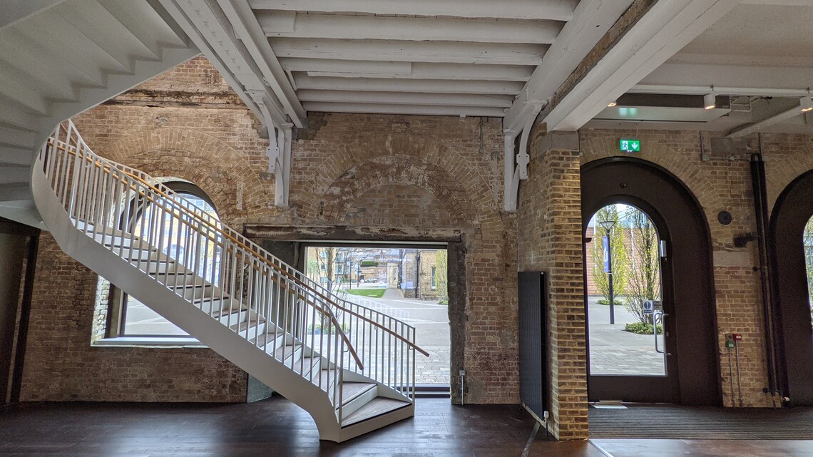 The foyer of Woolwich Works, with exposed brick walls, dark wooden flooring, large arched windows and doors, and a white metal staircase curving up to the left