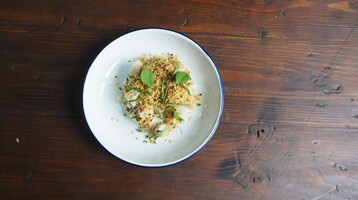 A white plate on a wooden table, of a lobster, cous cous and herb dish.