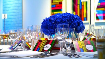 A photo of a table laid for an event in a room with colourful abstract pictures on the walls. Bright blue hydrangeas are centre pieces, and striped name cards match the wall art.