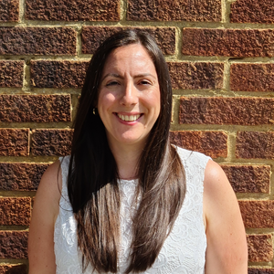 Ginene is a white woman with long brown hair, wearing a white sleeveless top. She is standing in front of a brick wall in the sunshine and smiling.