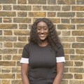 Justicia is a young black woman with long dark wavy hair, stood smiling in front of a brick wall.